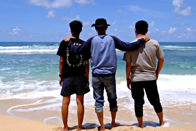 Three men standing together on the beach facing the ocean. Image by hartono subagio from Pixabay.